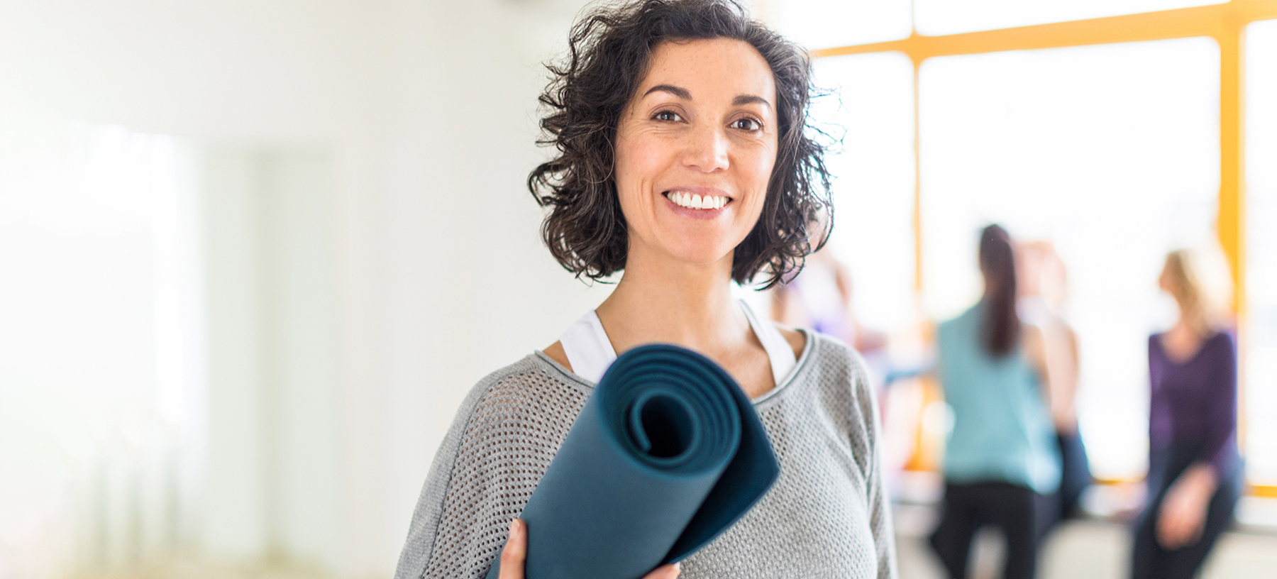 Smiling middle-aged woman holding a yoga mat after a wellness class—promoting active rehabilitation and mobility support for adult conditions.